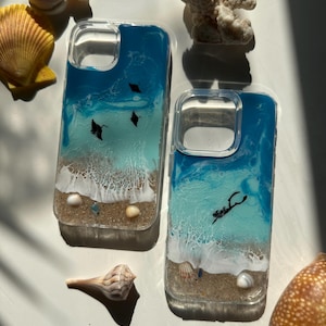 May include: Two clear phone cases with a beach scene design. The cases feature a blue and white ocean design with black manta rays and a diver. Seashells and coral are arranged around the cases.
