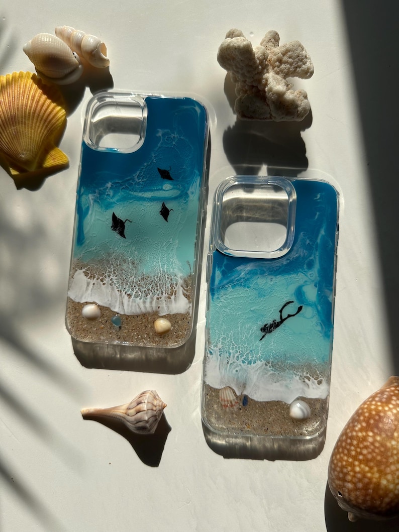May include: Two clear phone cases with a beach scene design. The cases feature a blue and white ocean design with black manta rays and a diver. Seashells and coral are arranged around the cases.