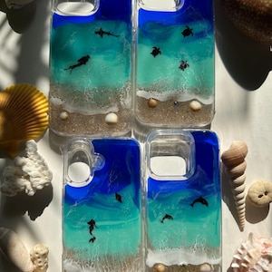 May include: Four clear phone cases with ocean-themed designs. Each case features a blue and turquoise resin ocean scene with sand, seashells, and small sea creatures like sharks, turtles, and dolphins. The cases are surrounded by seashells.