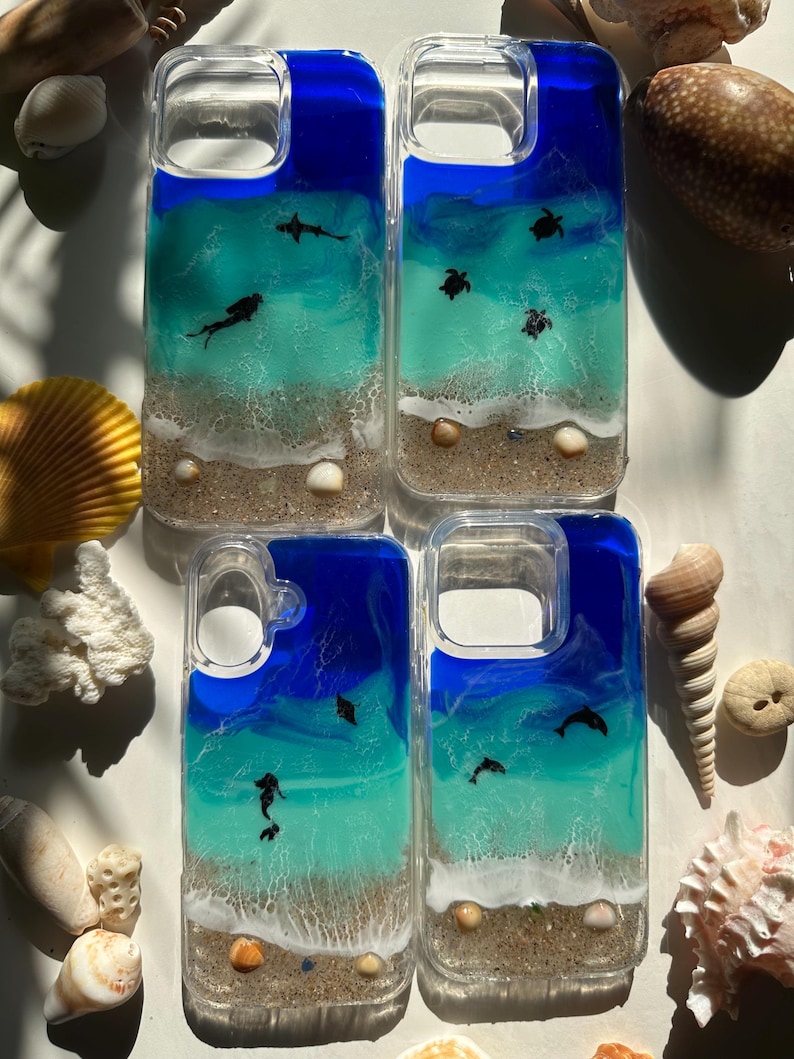 May include: Four clear phone cases with ocean-themed designs. Each case features a blue and turquoise resin ocean scene with sand, seashells, and small sea creatures like sharks, turtles, and dolphins. The cases are surrounded by seashells.