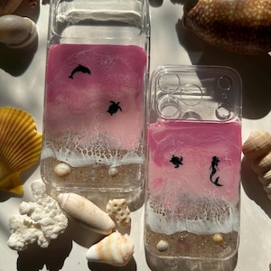May include: Clear phone cases with a pink and white ocean-themed design. One case features a dolphin and turtle, the other a turtle and mermaid. The cases have a sandy beach and ocean wave design with seashells.