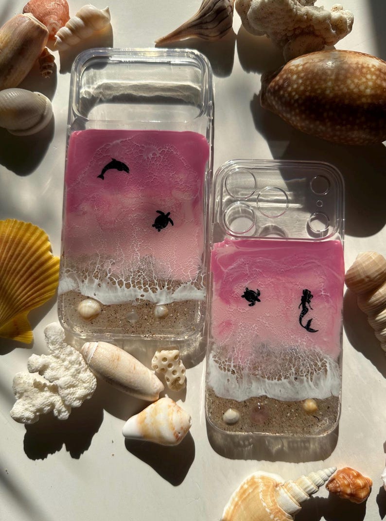 May include: Clear phone cases with a pink and white ocean-themed design. One case features a dolphin and turtle, the other a turtle and mermaid. The cases have a sandy beach and ocean wave design with seashells.