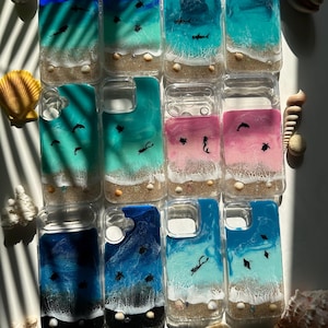 May include: A collection of clear phone cases with ocean-themed designs. Each case features a unique seascape with blue, teal, and pink resin, sandy beaches, and tiny sea creatures. Some cases include seashells.