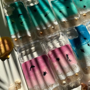 May include: Clear phone cases with ocean-themed designs. The cases feature a beach scene with sand, seashells, and marine life like dolphins, turtles, and mermaids. The ocean colors range from teal to pink and blue.