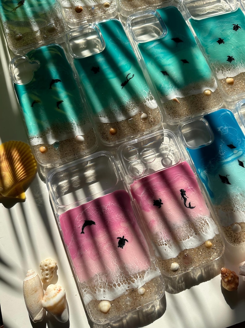 May include: Clear phone cases with ocean-themed designs. The cases feature a beach scene with sand, seashells, and marine life like dolphins, turtles, and mermaids. The ocean colors range from teal to pink and blue.
