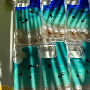 May include: Clear phone cases with ocean-themed designs. Each case features a blue and turquoise ocean scene with sand, seashells, and small black silhouettes of marine life. The cases are arranged on a white surface.