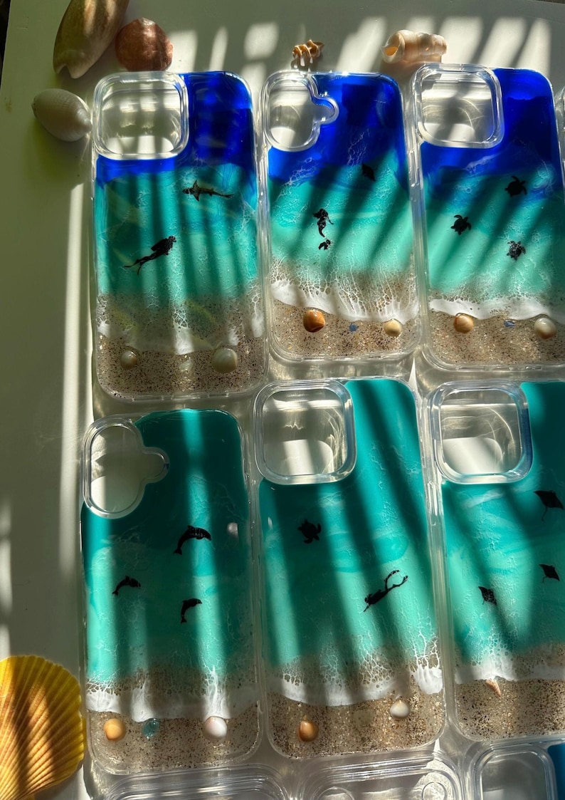 May include: Clear phone cases with ocean-themed designs. Each case features a blue and turquoise ocean scene with sand, seashells, and small black silhouettes of marine life. The cases are arranged on a white surface.