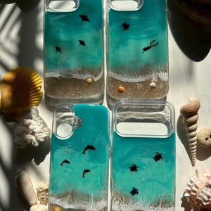 May include: Four clear phone cases with ocean-themed designs. Each case features a turquoise resin ocean scene with white waves, sand, and small sea creatures like stingrays, turtles, and dolphins. Small seashells and sand are visible at the bottom.