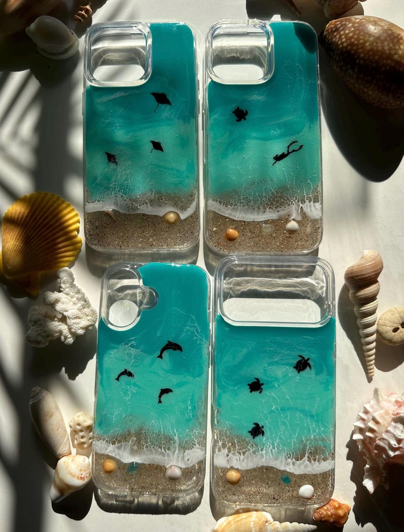 May include: Four clear phone cases with ocean-themed designs. Each case features a turquoise resin ocean scene with white waves, sand, and small sea creatures like stingrays, turtles, and dolphins. Small seashells and sand are visible at the bottom.