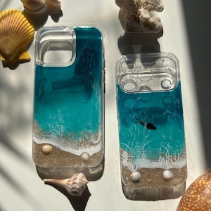 May include: Two clear phone cases with a beach-themed design. The cases feature layers of blue and turquoise resin, resembling ocean waves, with a sandy base and small seashells. A black fish silhouette is visible in one case.