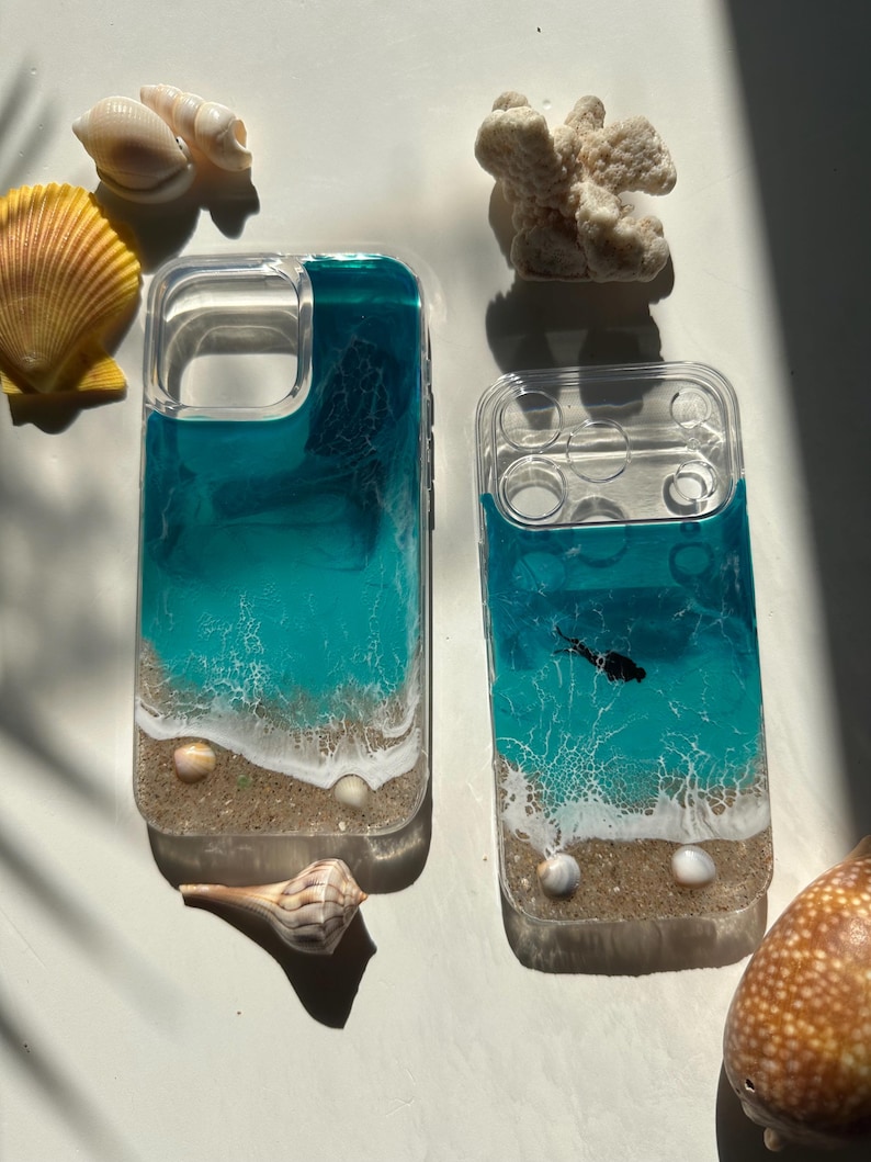 May include: Two clear phone cases with a beach-themed design. The cases feature layers of blue and turquoise resin, resembling ocean waves, with a sandy base and small seashells. A black fish silhouette is visible in one case.