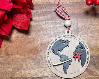 May include: Wooden Christmas ornament shaped like a globe with a red airplane flying over it. The ornament has the text "Cheers to another year of new destinations and unforgettable journeys".