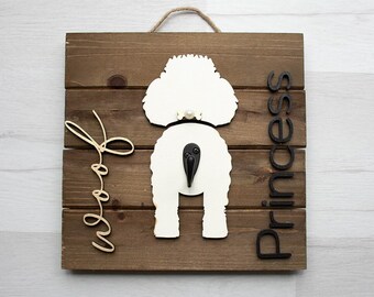 May include: A wooden sign with a white silhouette of a dog's rear end and the word "Princess" in black letters. The word "Woof" is written in cursive in gold letters on the left side of the sign.