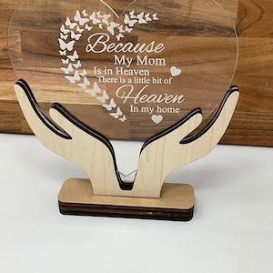May include: A clear acrylic heart-shaped memorial plaque with the text "Because My Mom Is in Heaven There is a little bit of Heaven In my home." The plaque is supported by a wooden base and hands design, with butterfly accents.