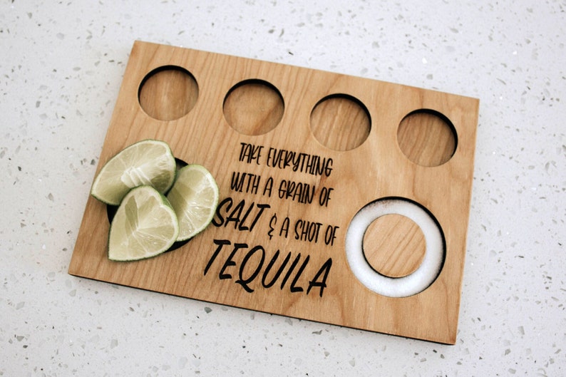 May include: Wooden tequila serving board with four circular cutouts for shot glasses, a central salt holder, and lime wedges. The board is engraved with the phrase "Take everything with a grain of salt & a shot of tequila."