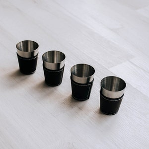 May include: Four shot glasses with stainless steel interiors and black leather exteriors. The leather is stitched with white thread. The shot glasses are arranged in a row on a light-colored wooden surface.