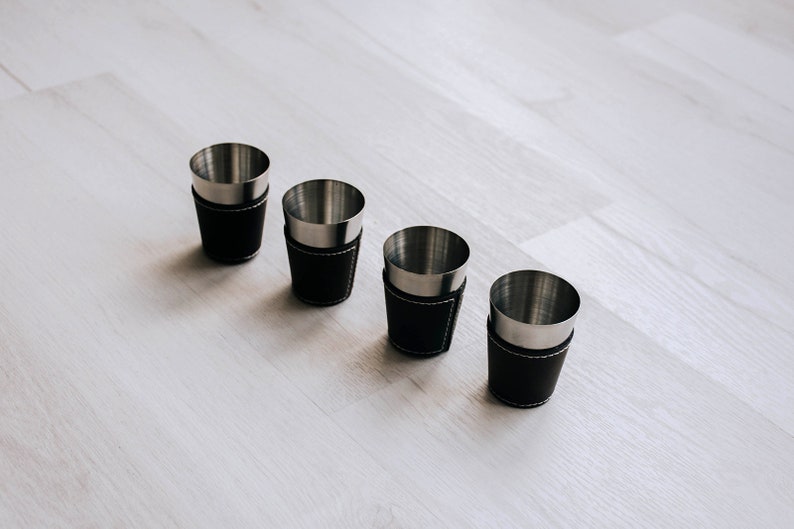 May include: Four shot glasses with stainless steel interiors and black leather exteriors. The leather is stitched with white thread. The shot glasses are arranged in a row on a light-colored wooden surface.