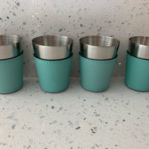 May include: Four shot glasses with stainless steel interiors and teal leatherette sleeves. The sleeves are stitched with black thread. The shot glasses are arranged in a row, offering a stylish presentation for beverages.