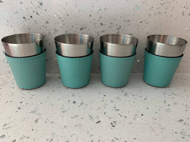 May include: Four shot glasses with stainless steel interiors and teal leatherette sleeves. The sleeves are stitched with black thread. The shot glasses are arranged in a row, offering a stylish presentation for beverages.
