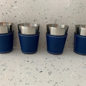 May include: Four stainless steel shot glasses with a navy blue leatherette sleeve and white stitching. The shot glasses have a silver rim and a tapered design. The set is displayed on a speckled white surface.