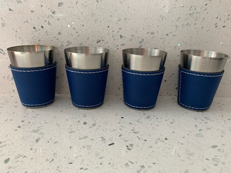 May include: Four stainless steel shot glasses with a navy blue leatherette sleeve and white stitching. The shot glasses have a silver rim and a tapered design. The set is displayed on a speckled white surface.