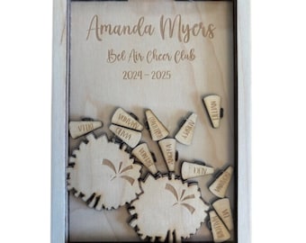 May include: A wooden box with a clear lid, containing a cheerleading pom-pom and small wooden megaphones with names engraved on them. The box is personalized with the name "Amanda Myers", "Bel Air Cheer Club", and the years "2024-2025".
