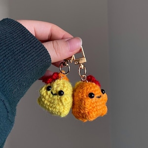 May include: Two handmade crochet keychains, one yellow and one orange, each with a red crest and black eyes. The keychains are attached to a gold-colored clasp. The crochet work is visible and detailed.