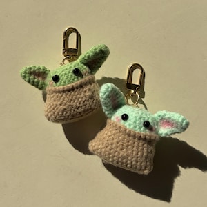 May include: Two handmade crochet keychains featuring a character with large ears, black eyes, and a tan robe. The keychains have gold-colored clasps. The character's ears are green with pink accents. The robe is tan.