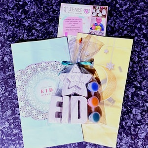 May include: A craft kit for children to paint plaster decorations. The kit includes a star, the word "Eid" and four bottles of paint. The kit is packaged in a clear plastic bag with a yellow and blue ribbon. The bag is sitting on top of a light blue envelope with a decorative pattern and the words "Eid Mubarak" in Arabic.