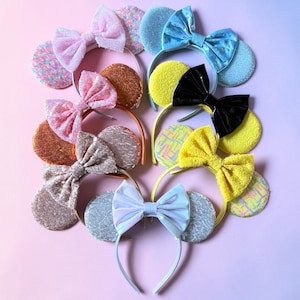 May include: A collection of seven headbands featuring circular ears and large bows. The headbands include various textures and colours such as pink sequins, metallic copper, silver glitter, light blue satin, black patent, and bright yellow fabric.