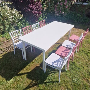 May include: A white rectangular table with six white and pink chairs. The table and chairs are set up outdoors on a grassy lawn.