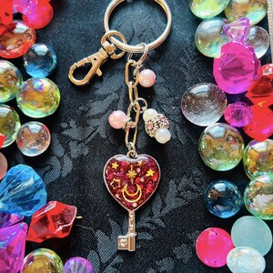 May include: A silver key charm with a red heart-shaped keyhole. The keyhole is filled with glitter and has a crescent moon and stars. The charm is attached to a silver chain and a silver key ring.