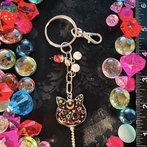 May include: A black cat charm keychain with a crescent moon and stars. The charm is attached to a silver chain with a silver ring and a silver clasp. The keychain is surrounded by colorful glass gems.