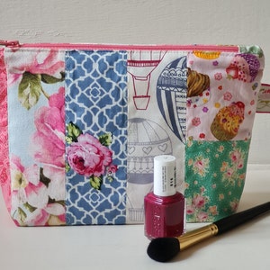 Peut inclure: Une pochette zipp&eacute;e en tissu patchwork avec un imprim&eacute; floral, g&eacute;om&eacute;trique et de montgolfi&egrave;res. La pochette est ouverte et un flacon de vernis &agrave; ongles et un pinceau &agrave; maquillage sont devant elle.