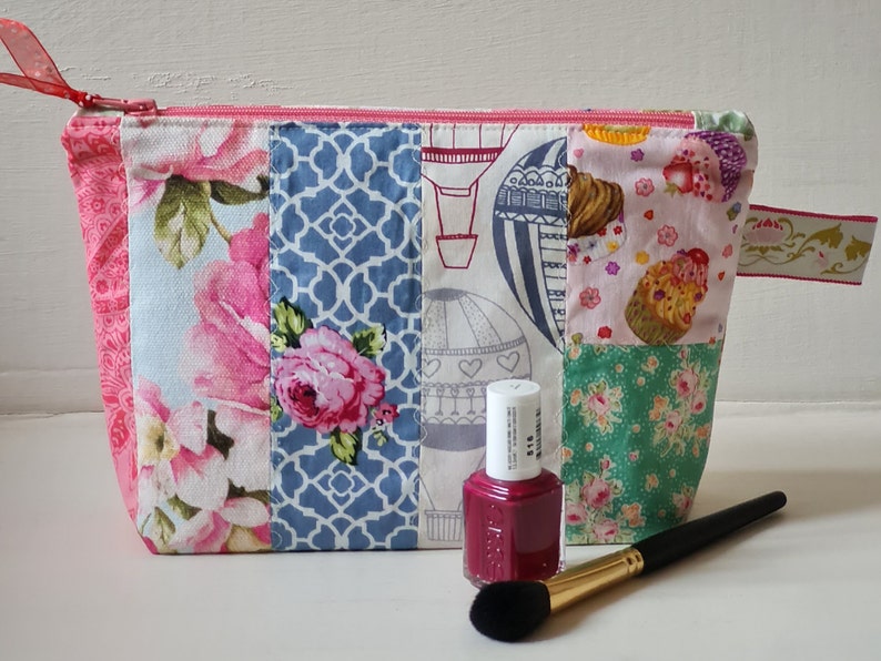 Peut inclure: Une pochette zipp&eacute;e en tissu patchwork avec un imprim&eacute; floral, g&eacute;om&eacute;trique et de montgolfi&egrave;res. La pochette est ouverte et un flacon de vernis &agrave; ongles et un pinceau &agrave; maquillage sont devant elle.