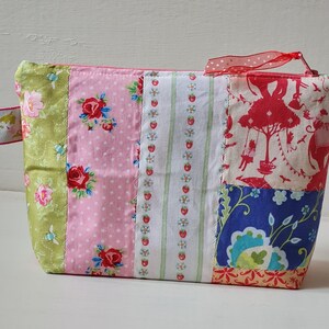 Peut inclure: Une pochette zipp&eacute;e en tissu patchwork avec un motif floral vert, rose, blanc et bleu. La pochette est dot&eacute;e d'un ruban rouge attach&eacute; &agrave; la fermeture &eacute;clair.