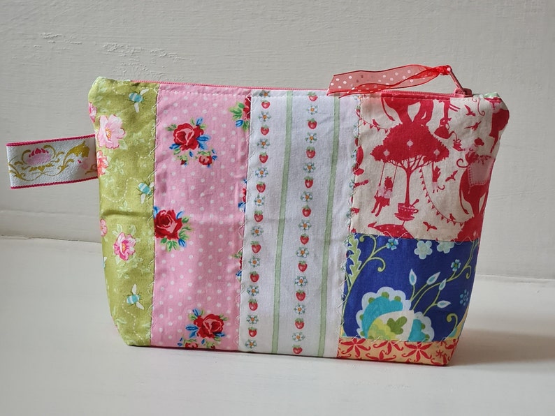 Peut inclure: Une pochette zipp&eacute;e en tissu patchwork avec un motif floral vert, rose, blanc et bleu. La pochette est dot&eacute;e d'un ruban rouge attach&eacute; &agrave; la fermeture &eacute;clair.