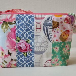 Könnte beinhalten: Eine Patchwork-Kosmetiktasche mit einem rosafarbenen Blumendesign, einem blauen geometrischen Muster und einem Heißluftballon-Druck. Die Tasche hat ein rosafarbenes Band mit weißen Punkten.