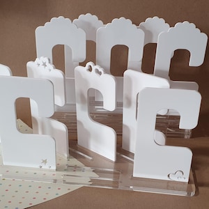 Może przedstawiać: Białe plastikowe wieszaki z wyciętym wzorem do zawieszania odzieży lub akcesoriów. Wieszaki są ułożone na przezroczystym stojaku akrylowym.