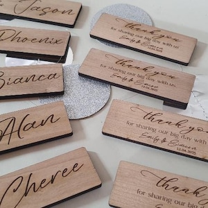 May include: Wooden place cards with names and a floral design. The cards are engraved with the names "Jason", "Phoenix", "Bianca", "Han", "Cheree" and the text "Thank you for sharing our big day with us Emily & Bernard 12.04.2023".