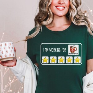 May include: A green t-shirt with a white graphic that says "I am working for winter break" with a rating of 5 stars and a red mug with a marshmallow on top.