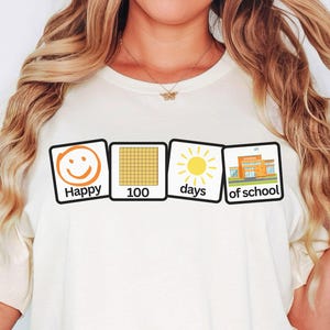 May include: Cream-colored t-shirt featuring four square graphics. The graphics include a smiling face with the word "Happy", a grid with the number "100", a sun with the word "days", and a school building with the words "of school".