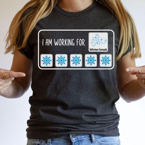 May include: Dark gray t-shirt with a white rectangular graphic. The graphic reads "I AM WORKING FOR" above five blue snowflake icons and a "Winter break" icon. The shirt is being pointed at by two hands.