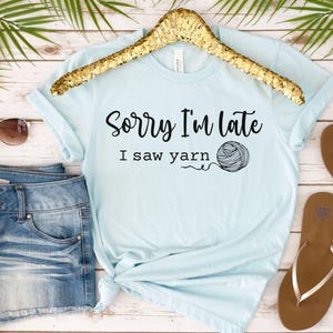 Op de afbeelding: Lichtblauw T-shirt met de tekst "Sorry, ik ben te laat, ik zag garen" en een zwart-witte illustratie van een bol garen.