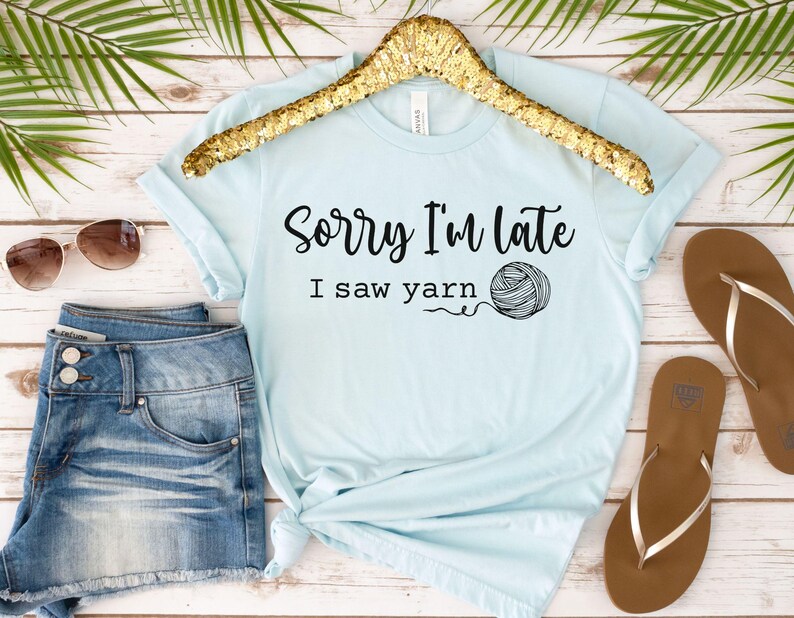 Op de afbeelding: Lichtblauw T-shirt met de tekst "Sorry, ik ben te laat, ik zag garen" en een zwart-witte illustratie van een bol garen.