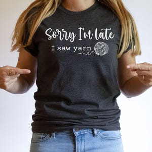Op de afbeelding: Een donkergrijs T-shirt met de tekst "Sorry, ik ben te laat, ik zag garen" en een witte omtrek van een bol garen.