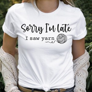 Op de afbeelding: Wit T-shirt met zwarte tekst die "Sorry, ik ben te laat, ik zag garen" zegt en een zwart-witte illustratie van een bol garen.