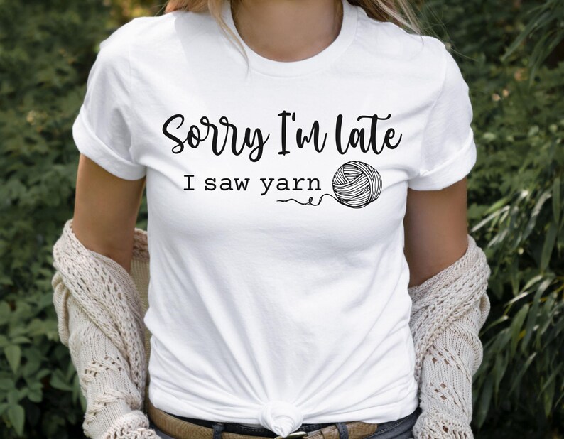 Op de afbeelding: Wit T-shirt met zwarte tekst die "Sorry, ik ben te laat, ik zag garen" zegt en een zwart-witte illustratie van een bol garen.
