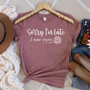 Op de afbeelding: Een mauve t-shirt met de tekst "Sorry, ik ben te laat, ik zag garen" en een witte illustratie van een bol garen.