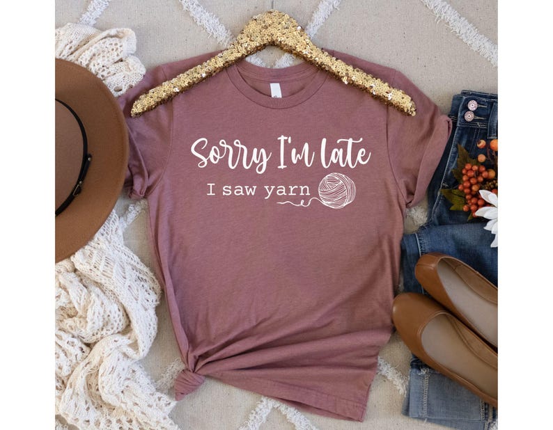 Op de afbeelding: Een mauve t-shirt met de tekst "Sorry, ik ben te laat, ik zag garen" en een witte illustratie van een bol garen.